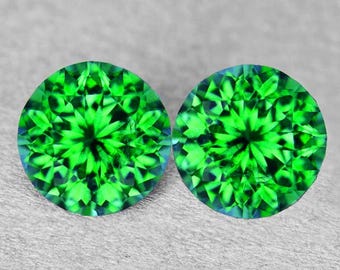 May include: Two round, faceted gemstones with a vibrant, bright green color. The gems have a complex, starburst-like pattern of facets, creating a sparkling effect. The background is a neutral, light gray.