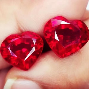 Può includere: Due pietre preziose di rubino a forma di cuore, ciascuna con un colore rosso intenso e un taglio sfaccettato.