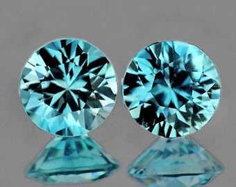 Puede incluir: Dos gemas redondas y facetadas de un brillante color azul claro. Las gemas se muestran sobre una superficie reflectante, mostrando sus intrincados cortes y reflejos internos. Las gemas son de un tono azul vibrante y llamativo.