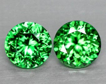 May include: Two round, faceted gemstones with a vibrant, emerald green color. The gems have a brilliant cut, reflecting light to create a sparkling effect. The gemstones are displayed against a neutral gray background.