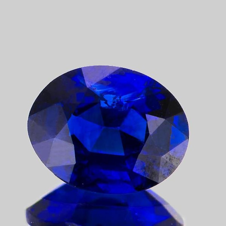May include: A faceted, oval-shaped, deep blue gemstone. The gem has a high degree of clarity and brilliance, with multiple facets reflecting light. The stone is set against a light gray background, with a slight reflection visible below.
