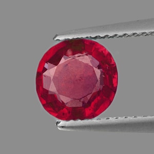 Könnte beinhalten: Ein runder, roter Edelstein mit facettiertem Schliff. Der Edelstein wird in einer Pinzette gehalten.