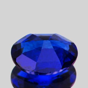 May include: A faceted, oval-shaped, deep blue gemstone. The gem is highly reflective, with multiple facets catching the light. The stone is set against a light gray background, with its reflection visible below.