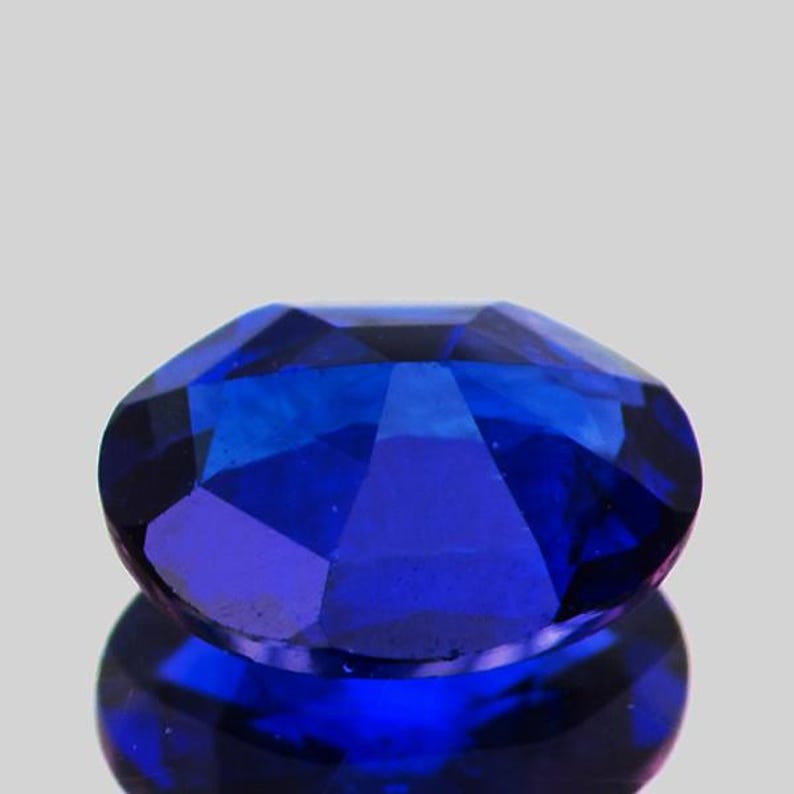 May include: A faceted, oval-shaped, deep blue gemstone. The gem is highly reflective, with multiple facets catching the light. The stone is set against a light gray background, with its reflection visible below.