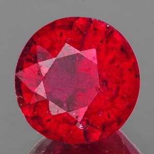 May include: A faceted, round-cut ruby gemstone. The gem is a deep, saturated red color with internal inclusions and a star-like pattern in the center. The stone is polished and reflects light.