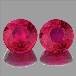 Puede incluir: Dos piedras preciosas de rubí redondas y facetadas de color rojo intenso. Las piedras preciosas están colocadas sobre un fondo gris.