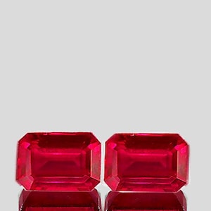 Peut inclure: Deux pierres précieuses de couleur rouge vif taillées en émeraude, probablement des rubis, sont présentées sur un fond blanc. Les pierres précieuses sont placées côte à côte, mettant en valeur leur brillance et leur clarté.