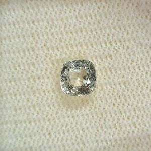 May include: A light grey cushion-cut gemstone. The square-shaped gem has rounded corners and multiple facets, reflecting light. It is set against a white, textured background, accentuating its clarity and sparkle. A piece of jewellery.
