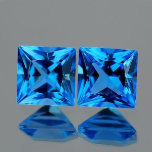 May include: Two square-shaped, bright blue topaz gemstones with a faceted cut.