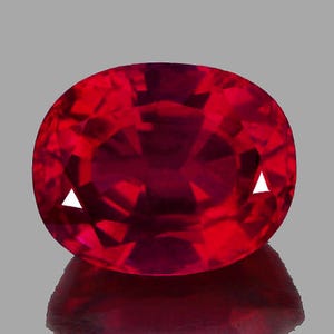 May include: A deep red oval-shaped gemstone with a faceted cut. The gemstone is set against a gray background.