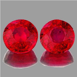 May include: Two round, faceted, deep red gemstones with a brilliant shine. The gems are displayed on a reflective surface, showcasing their colour and cut. The gemstones are a vibrant shade of red, with visible internal structures.
