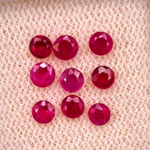 May include: Eight round, faceted rubies with a deep red colour and brilliant cut. Each gemstone displays a symmetrical circular shape with multiple facets that reflect light, arranged in a loose cluster on a textured, light-coloured fabric surface.