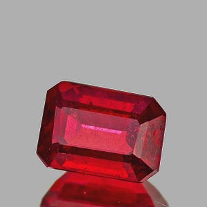 May include: A faceted, rectangular ruby gemstone with a deep red hue. The gem has an emerald cut with beveled edges, reflecting light and showcasing its clarity. The ruby is set against a neutral background, highlighting its vibrant color and brilliance.