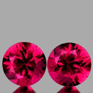 May include: Two round-cut gemstones with a vibrant, deep red color and a faceted surface. Each stone displays a symmetrical pattern of geometric cuts that reflect light, creating a brilliant, sparkling appearance against a neutral background.