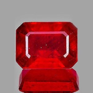 May include: A deep red gemstone with a rectangular emerald cut. The gemstone is faceted and has a high polish.