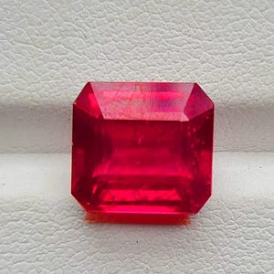 May include: A faceted, rectangular-cut, vibrant red gemstone. The gem has a clear, polished surface with multiple facets reflecting light. The gemstone is set against a textured, off-white background. This is a close-up of a ruby.