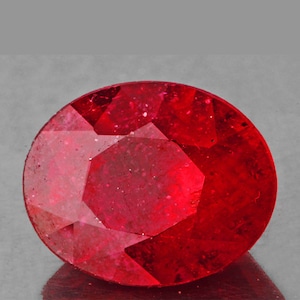 May include: A deep red oval ruby gemstone with a faceted cut. The gemstone is set against a gray background.