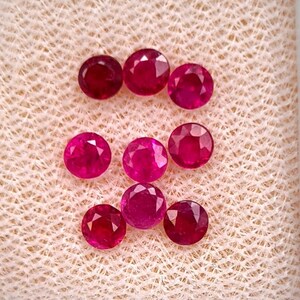 May include: Eight round-cut, vibrant red rubies arranged in a cluster. Each gemstone features a faceted surface that reflects light, showcasing a deep crimson hue and a circular shape. The stones are displayed together, highlighting their consistent color.