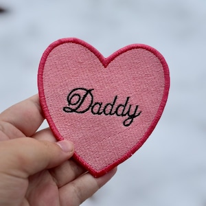 May include: Pink heart-shaped embroidered patch with the word "Daddy" in black script. The patch has a pink border and a textured appearance, suitable for clothing or accessories.
