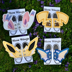 Könnte beinhalten: Vier Paar Schuhflügel in verschiedenen Designs, darunter gelbe Blitz, blaue Fledermaus und rosa und lila Flügelformen. Jedes Paar ist an einer Karte mit dem Text "Shoe Wings" und einer Schuhkontur befestigt. Die Flügel sind aus Stoff und haben Metallnieten.