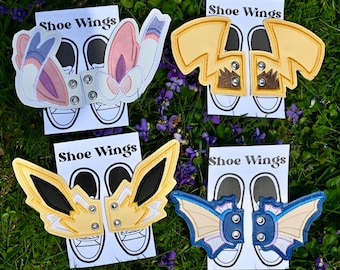 May include: Four sets of embroidered shoe wings attached to white display cards labeled Shoe Wings. The designs include pink and white rabbit ears, yellow lightning bolt shapes, yellow and black pointed wings, and blue and cream bat-like wings.