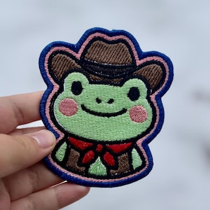 May include: Embroidered patch featuring a cartoon frog wearing a brown cowboy hat and a red neckerchief. The frog has a green body, pink cheeks, and a smiling expression. The patch is outlined in blue and pink.