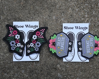 May include: Two pairs of shoe wings. One pair is black with pink and white flowers and the other pair is gray with a tombstone design that says "Dead Inside".