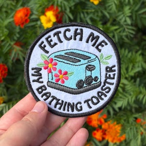 May include: A circular embroidered patch with a white background and a black border. It features a light blue toaster illustration with pink flowers and black text that reads, "FETCH ME MY BATHING TOASTER" in a bold, black, sans-serif font.