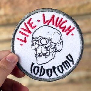 May include: Round white patch with a black and white skull design. The words "LIVE. LAUGH." are embroidered in red above the skull, and the word "lobotomy" is below the skull in black.