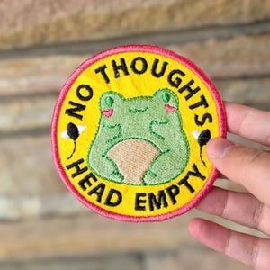 May include: Embroidered patch with a yellow background and a pink border. The patch features a green frog with the text "NO THOUGHTS HEAD EMPTY" in black. Two small black and white details are on either side of the frog.
