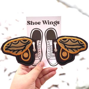 Może przedstawiać: Haftowane skrzydła do butów w kolorach brązu, beżu i złota. Skrzydła mają kształt skrzydeł motyla i mają szczegółowy wzór. Tekst "Shoe Wings" jest wydrukowany na białej karcie.