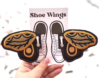 May include: Embroidered shoe wings in brown, tan, and gold. The wings are shaped like butterfly wings and have a detailed design. The text "Shoe Wings" is printed on a white card behind the wings.