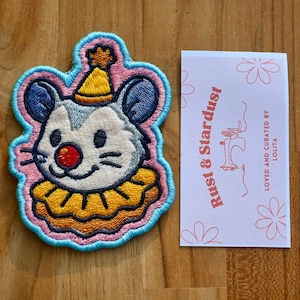 May include: Embroidered patch featuring a cartoon mouse wearing a party hat and ruffled collar. The patch has a pink and blue border. A card with the text "Rust & Stardust" and "Loved and curated by Lolita" is next to the patch.