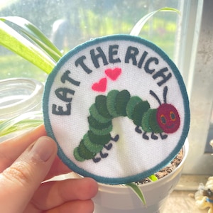 May include: A white, round embroidered patch with a green caterpillar, red heart shapes, and the text "Eat the Rich".