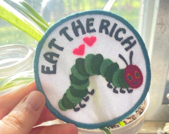 May include: A white, round embroidered patch with a green caterpillar, red heart shapes, and the text "Eat the Rich".