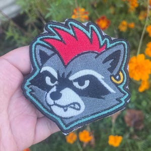 May include: Embroidered patch featuring a stylized raccoon with a red mohawk, a gold earring, and a teal outline. The raccoon has a gray face with black markings and a stern expression. This patch is suitable for jackets, bags, or other fabric items.