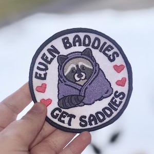 May include: Round patch with a raccoon wearing a purple hooded blanket. The patch has the text "EVEN BADDIES GET SADDIES" in a circular pattern around the raccoon. Red hearts are scattered around the raccoon.