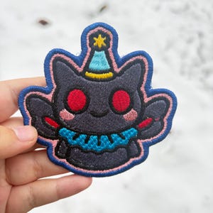 May include: Embroidered patch featuring a dark gray cartoon character with red eyes, a blue party hat, and a blue collar. The patch has a blue border with pink accents. The character has a smiling expression.