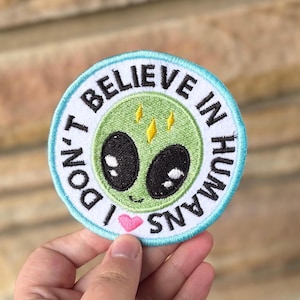 Op de afbeelding: Een ronde geborduurde patch met een lichtblauwe rand en de tekst "I DON'T BELIEVE IN HUMANS". De patch toont een groen alien gezicht met grote zwarte ogen, een kleine glimlach en een roze hart.