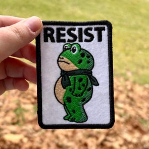 Puede incluir: Parche bordado con una rana verde con lunares negros y una bufanda negra, sobre un fondo blanco. La palabra "RESIST" está impresa en negro sobre la rana. El parche tiene un borde negro.