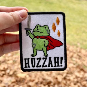 May include: Embroidered patch featuring a green frog wearing a red cape and holding a sword. The patch has the word "Huzzah!" in bold letters. The frog is on a white background with a black border and orange diamond shapes.