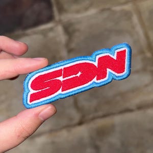 May include: Embroidered patch with the letters "SDN" in red, outlined in white, and bordered by a light blue stitching. The patch is held between fingers against a neutral background.