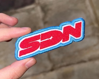 May include: Embroidered patch with the letters "SDN" in red, outlined in white, and bordered by a light blue stitching. The patch is held between fingers against a neutral background.