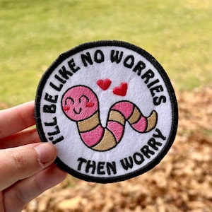 May include: Round embroidered patch with a smiling pink worm, two red hearts, and the text "I'll be like no worries then worry" in black. The patch has a white background and a black border. The worm has a gold and pink striped body.