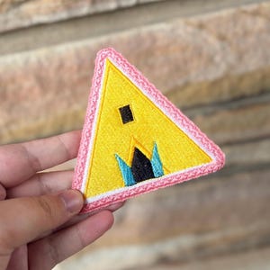 May include: A yellow triangular patch with a pink border. The patch features a black square and a stylized design with blue and black elements. The patch is held in a hand.