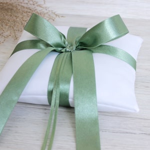 Può includere: Un cuscino per anelli in raso bianco con un nastro in raso verde salvia legato a fiocco. Il nastro ha code lunghe che pendono da entrambi i lati del cuscino.