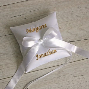 May include: A white satin ring pillow with gold embroidered names "Margaret" and "Jonathan". The pillow is square, tied with a white satin bow and ribbon.