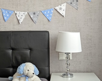 May include: A white plush toy dog with blue ears and a blue bow sits on a black leather headboard. A white lamp with a clear glass base and a white shade sits on a white nightstand. A blue and gray star garland hangs above the headboard.