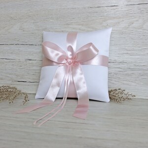 May include: A white satin ring pillow with a large pink satin bow and long tails. The pillow is on a light wood background.