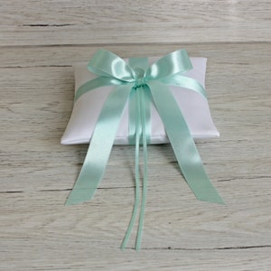 Può includere: Un cuscino per anelli in raso bianco con un fiocco in raso blu chiaro e code lunghe. Il cuscino è su uno sfondo in legno chiaro.